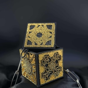 May include: Black box with gold beaded geometric designs on each side and the lid. The lid is slightly open, revealing the interior. A silver chain is attached to the box. The box is sitting on a black surface.