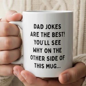 Op de afbeelding: Witte keramische mok met de tekst "DAD JOKES ARE THE BEST! YOU'LL SEE WHY ON THE OTHER SIDE OF THIS MUG..." in zwart. De mok wordt vastgehouden door een persoon die een crèmekleurige trui draagt.