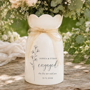 May include: A white ceramic vase with a scalloped rim and a sheer ribbon tied around the neck. The vase features a black floral line drawing and the text: SOPHIA & ETHAN, engaged, the day we said yes, 10.12.2026. It is filled with pale pink roses.