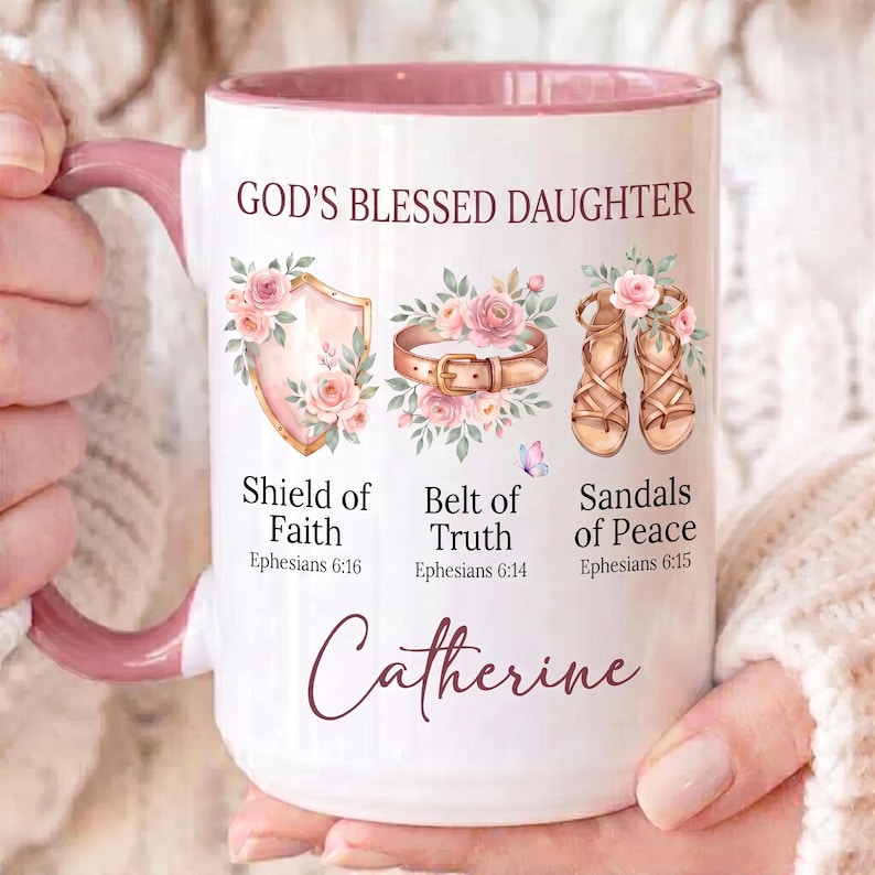 May include: White ceramic mug with a pink interior and handle, featuring the text "GOD'S BLESSED DAUGHTER." The mug is decorated with illustrations of a shield, belt, and sandals, along with the name "Catherine" in cursive. The mug is held by a person.