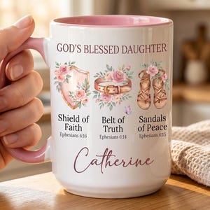 May include: White ceramic mug with a pink interior and handle. The mug features the text "GOD'S BLESSED DAUGHTER" and illustrations of a shield, belt, and sandals, along with the name "Catherine". Floral accents and script font add to the design.
