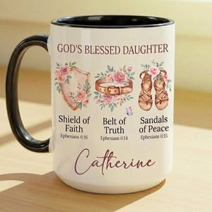 May include: White ceramic mug with a black handle and rim, featuring the text "GOD'S BLESSED DAUGHTER" and the name "Catherine". The mug is decorated with floral illustrations of a shield, belt, and sandals, with accompanying text from Ephesians.