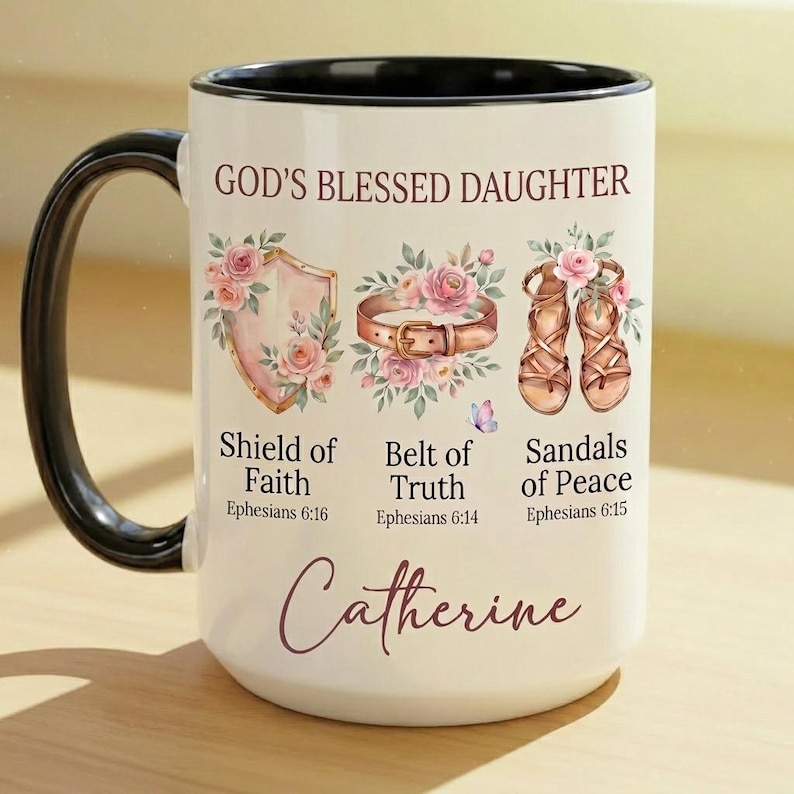 May include: White ceramic mug with a black handle and rim, featuring the text "GOD'S BLESSED DAUGHTER" and the name "Catherine". The mug is decorated with floral illustrations of a shield, belt, and sandals, with accompanying text from Ephesians.