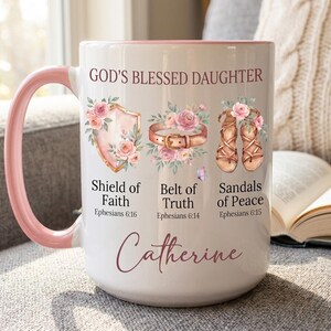 May include: White ceramic mug with a pink interior and handle. The mug features the text "GOD'S BLESSED DAUGHTER" and illustrations of a shield, belt, and sandals, each adorned with flowers. The name "Catherine" is written in cursive script.