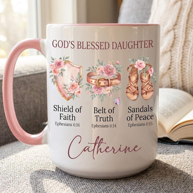 May include: White ceramic mug with a pink interior and handle. The mug features the text "GOD'S BLESSED DAUGHTER" and illustrations of a shield, belt, and sandals, each adorned with flowers. The name "Catherine" is written in cursive script.