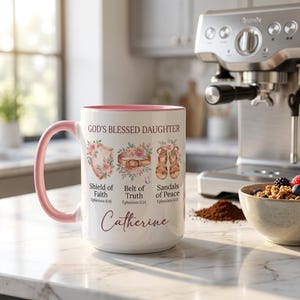 May include: White ceramic mug with a pink handle and rim, featuring the text "GOD'S BLESSED DAUGHTER" and floral illustrations. The mug includes the name "Catherine" and references to Ephesians. A coffee machine and a bowl of cereal are in the background.