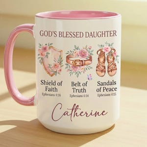 May include: A white ceramic mug with a pink handle and interior rim. The mug features the text "GOD'S BLESSED DAUGHTER" and illustrations of a shield, belt, and sandals, with the name "Catherine" below. Floral accents and script font.