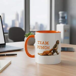 May include: White ceramic mug with an orange interior and handle. The mug features the text "TEAM PUNCH" in orange, along with an illustration of a monkey punching a cat. The mug is on a wooden desk.