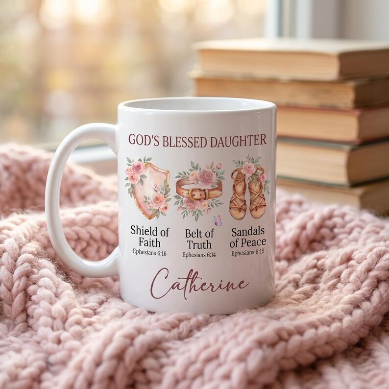 May include: White ceramic mug with the text "GOD'S BLESSED DAUGHTER" and the name "Catherine". The mug features illustrations of a shield, belt, and sandals, with floral accents. The text "Shield of Faith", "Belt of Truth", and "Sandals of Peace" are also printed on the mug.