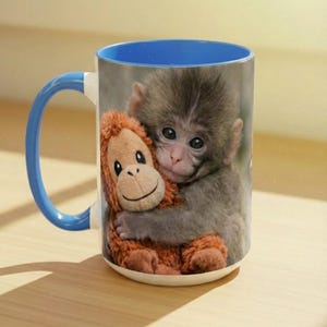 May include: White ceramic mug with a blue interior and handle. The mug features a print of a baby monkey hugging a stuffed orangutan. The monkey has grey fur and the stuffed animal is brown.