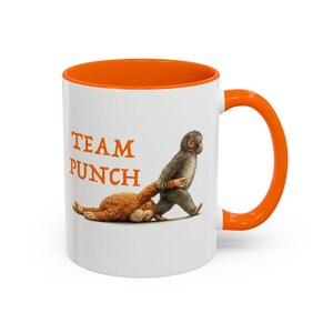 May include: White ceramic mug with an orange interior and handle. The mug features the text "TEAM PUNCH" in orange, along with an illustration of a monkey dragging a stuffed animal. A fun and playful design.