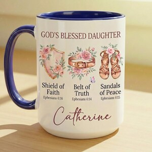 May include: White ceramic mug with a blue handle and interior rim. The mug features the text "GOD'S BLESSED DAUGHTER" and illustrations of a shield, belt, and sandals, with the name "Catherine" in script.