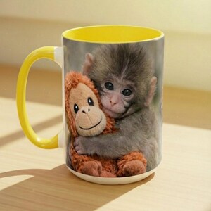 May include: A white ceramic mug with a yellow interior and handle. The mug features a smiling baby monkey hugging a stuffed orangutan. The monkey has grey fur, and the stuffed animal is brown.