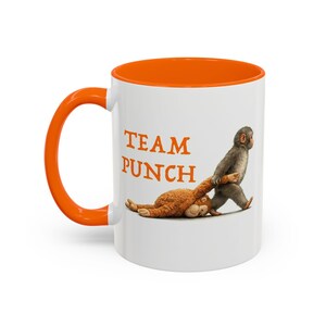May include: White ceramic mug with an orange interior and handle. The mug features the text "TEAM PUNCH" in orange letters, along with an illustration of a monkey dragging a stuffed animal. A fun and playful design.