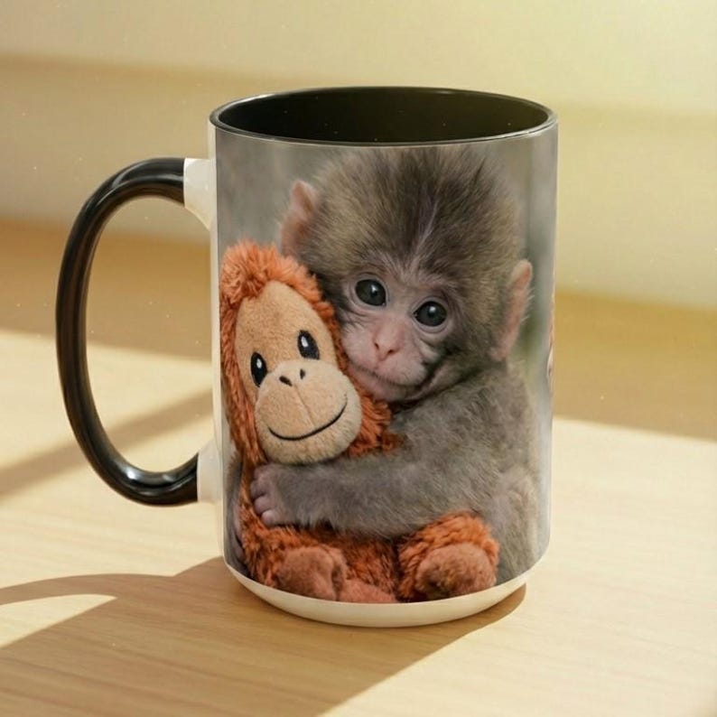 May include: White ceramic mug with a black handle and rim. The mug features a print of a baby monkey hugging a stuffed orangutan. The monkey has grey fur and the stuffed animal is orange. The mug is suitable for hot or cold drinks.