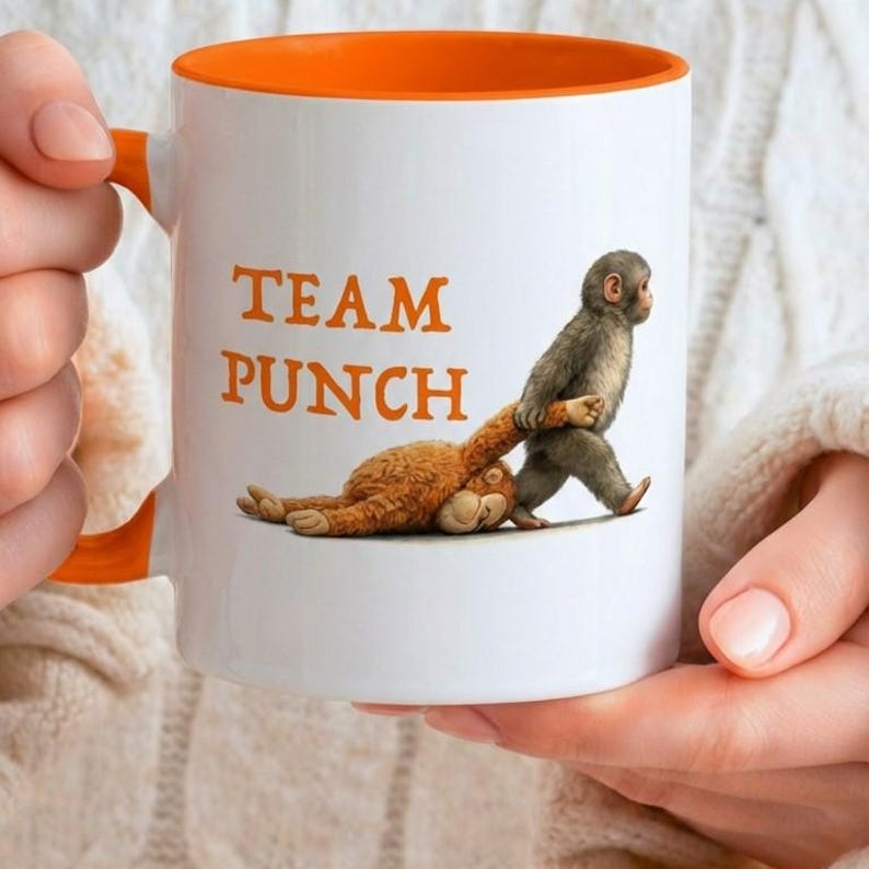 May include: White ceramic mug with an orange interior and handle. The mug features the text "TEAM PUNCH" in orange, along with an illustration of two monkeys. One monkey is pulling the other by the arm. The mug is held by a person.