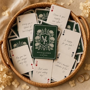 Peut inclure: Un ensemble de cartes à jouer personnalisées dans un panier en osier. Les cartes présentent un dos vert foncé avec un blason floral, les initiales ML et le texte Mitch & Lauren 09.26.2026. D'autres cartes affichent des vœux de mariage manuscrits.