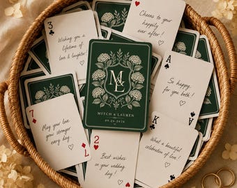 Può includere: Un set di carte da gioco personalizzate in un cesto intrecciato. Le carte presentano un retro verde scuro con uno stemma floreale, le iniziali ML e il testo Mitch & Lauren 09.26.2026. Altre carte mostrano auguri di matrimonio scritti a mano.