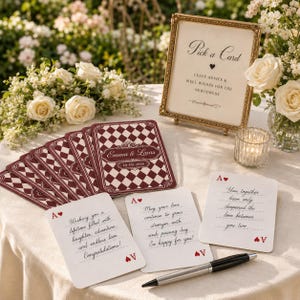 May include: A set of wedding advice cards featuring a maroon and white diamond pattern on the back with the text Emma & Lucas 10-05-2026. The front of the cards are white with red ace of hearts symbols and handwritten messages. A gold frame displays Pick a Card.
