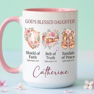May include: White ceramic mug with a pink handle and interior. The mug features the text "GOD'S BLESSED DAUGHTER" and illustrations of a shield, belt, and sandals, with the words "Shield of Faith," "Belt of Truth," and "Sandals of Peace." The name "Catherine" is also printed on the mug.