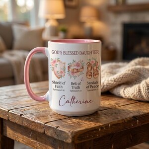 May include: White ceramic mug with a pink handle and interior rim. The mug features the text "GOD'S BLESSED DAUGHTER" and illustrations of a shield, belt, and sandals, along with the name "Catherine".