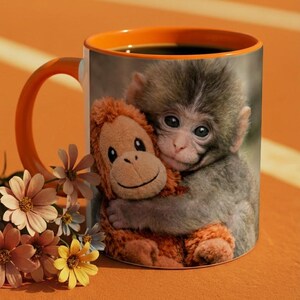 May include: A ceramic coffee mug with an orange handle and interior. The mug features a print of a baby monkey hugging a stuffed monkey. The mug is filled with a dark liquid, and flowers are arranged near the base.