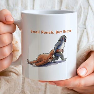 May include: White ceramic mug featuring an illustration of a small monkey punching a sloth. The text "Small Punch, But Brave" is printed in orange. The mug is held by a person.