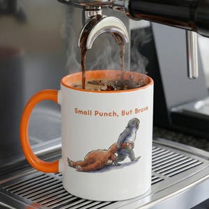 May include: A white ceramic mug with an orange handle and interior is filled with coffee from an espresso machine. The mug features a cartoon illustration of two creatures and the text "Small Punch, But Brave."