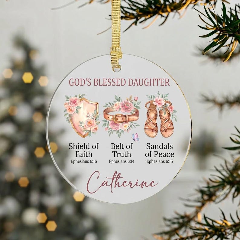 May include: A clear, round Christmas ornament with gold ribbon. The ornament features the text "GOD'S BLESSED DAUGHTER" and illustrations of a shield, belt, and sandals, each with floral accents. The name "Catherine" is written at the bottom.
