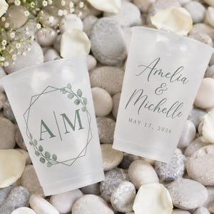 Op de afbeelding: Twee matte plastic bekers met ijs, gepersonaliseerd voor een bruiloft. Eén beker heeft de initialen "AM" in een geometrisch ontwerp met bladeren. De andere beker is beschreven met "Amelia & Michelle" en de datum "17 mei 2026".
