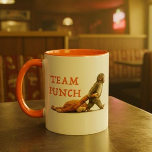 May include: White ceramic mug with an orange interior and handle. The mug features the text "TEAM PUNCH" in orange, along with an illustration of a monkey pulling another monkey. The mug is sitting on a table.