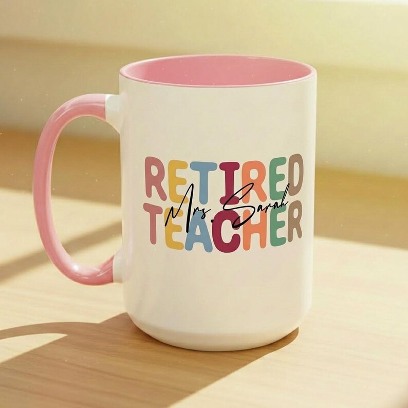 K&ouml;nnte beinhalten: Wei&szlig;er Keramikbecher mit rosa Innenraum und Henkel. Der Becher zeigt die Worte "RETIRED Mrs. Sarah TEACHER" in bunten Blockbuchstaben. Ein durchdachtes Geschenk f&uuml;r eine pensionierte Lehrerin.