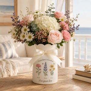 Könnte beinhalten: Eine weiße Keramikvase mit gewelltem Rand und einer cremefarbenen Schleife. Die Vase zeigt Blumenillustrationen, den Text "Grandma's Garden", "Emily", "Henry", "Lucy" und "Love grows here" und ist mit weißen Hortensien und rosa Rosen gefüllt.