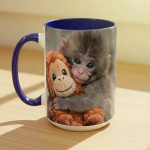 May include: White ceramic mug with a blue interior and handle. The mug features a smiling baby monkey hugging a stuffed orangutan. The monkey has grey fur and the stuffed animal is brown. Ideal for tea, coffee, or hot chocolate.