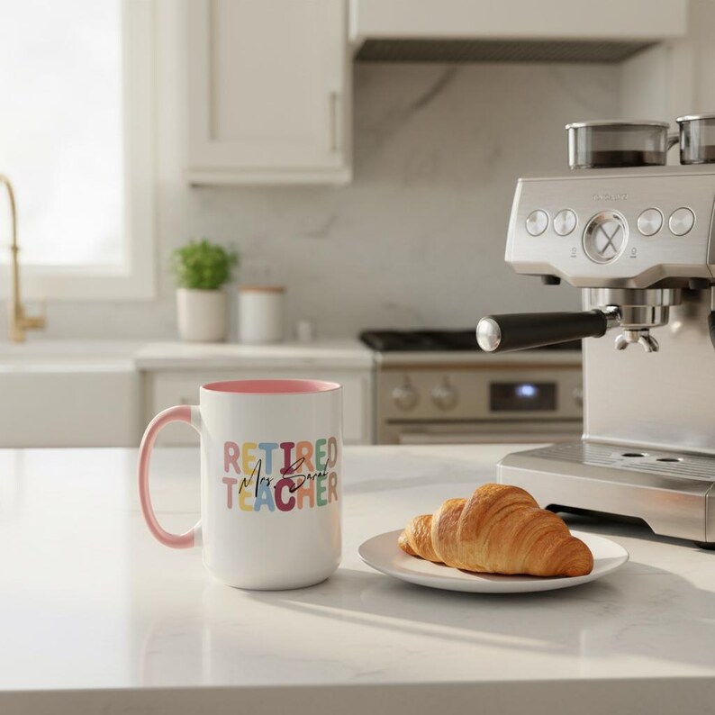 K&ouml;nnte beinhalten: Eine wei&szlig;e Keramiktasse mit rosa Innenraum und Griff. Die Tasse zeigt die Worte "RETIRED TEACHER" in bunten Blockbuchstaben. Ein Teller mit zwei Croissants steht neben der Tasse, in der N&auml;he einer Espressomaschine aus Edelstahl.