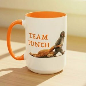 May include: White ceramic mug with an orange interior and handle. The mug features the text "TEAM PUNCH" in orange, along with an illustration of two monkeys. One monkey is pulling the other by the arm.