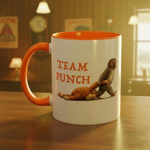 May include: White ceramic mug with an orange interior and handle. The mug features the text "TEAM PUNCH" in orange, along with an illustration of a monkey dragging a stuffed animal. The mug is on a wooden surface.