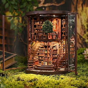 Op de afbeelding: Een gedetailleerd houten miniatuur diorama van een boekenkast, met meerdere niveaus van boekenplanken, een wenteltrap en een kleine boom. Het diorama heeft een warme, uitnodigende gloed van de binnenverlichting en een achtergrond van groen.