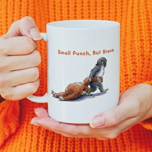 May include: White ceramic mug featuring a cartoon illustration of a small monkey punching a sloth. The text "Small Punch, But Brave" is printed above the illustration in brown. The mug has a curved handle.