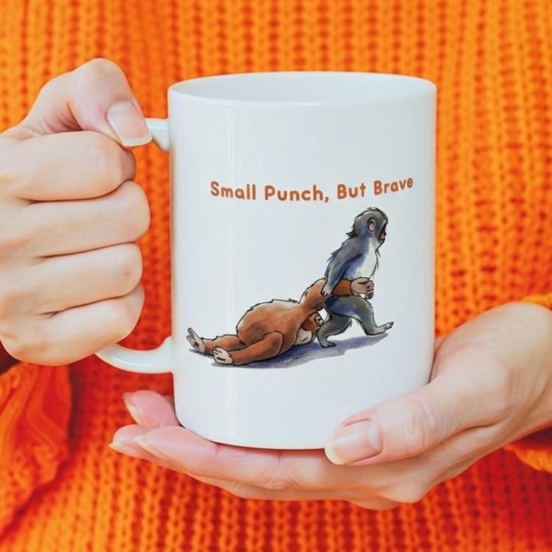 May include: White ceramic mug featuring a cartoon illustration of a small monkey punching a sloth. The text "Small Punch, But Brave" is printed above the illustration in brown. The mug has a curved handle.
