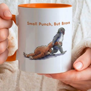 May include: White ceramic mug with an orange interior and handle. The mug features an illustration of two cartoon monkeys, one brown and one gray, with the text "Small Punch, But Brave" in orange letters.