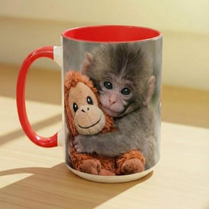 May include: White ceramic mug with a red interior and handle. The mug features a print of a baby monkey hugging a stuffed orangutan. The monkey has grey fur and the stuffed animal is brown. The mug is on a light brown surface.