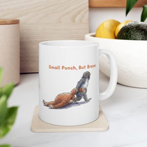 May include: White ceramic mug featuring an illustration of two cartoon monkeys. One monkey is pulling the other. The text "Small Punch, But Brave" is printed in orange. The mug is on a beige coaster.