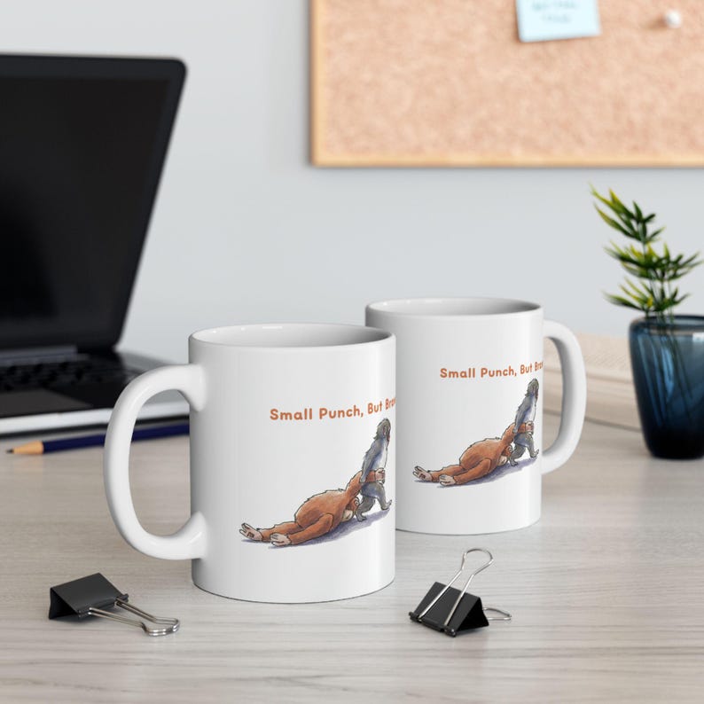 May include: Two white ceramic mugs with a humorous illustration of two figures in a wrestling pose. The mugs have the text "Small Punch, But Big" in orange. The mugs are on a light-colored surface.