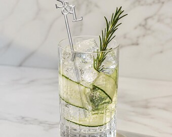 Può includere: Un bicchiere trasparente riempito con cubetti di ghiaccio, fettine sottili di cetriolo e un rametto di rosmarino verde. Un agitatore per cocktail in metallo argentato con le lettere S e L in carattere serif è inserito nella bevanda.