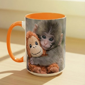 May include: A ceramic mug with an orange handle and interior. The mug features a print of a baby monkey hugging a stuffed animal. The stuffed animal is brown and the monkey is gray. The mug is on a light-colored surface.
