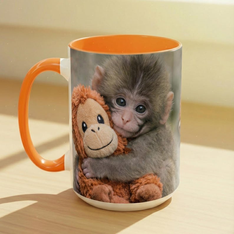 May include: A ceramic mug with an orange handle and interior. The mug features a print of a baby monkey hugging a stuffed toy. The toy is brown and the monkey is grey. The mug is on a light-coloured surface.