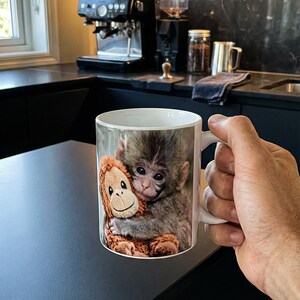 May include: White ceramic mug featuring a baby monkey hugging a stuffed orangutan. The mug has a light background and is held by a person. The mug is likely for coffee or tea.