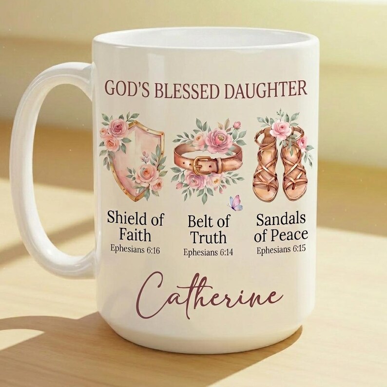May include: White ceramic mug with the text "GOD'S BLESSED DAUGHTER" and the name "Catherine". The mug features illustrations of a shield, belt, and sandals, each adorned with floral arrangements and associated Bible verses.
