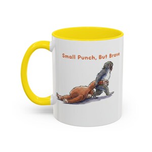May include: White ceramic mug with a yellow interior and handle. The mug features an illustration of two cartoon animals, one brown and one gray, in a playful wrestling pose. The text "Small Punch, But Brave" is printed on the mug.